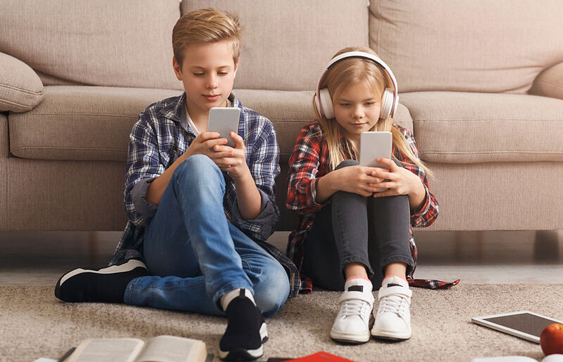 Zwei Kinder sitzen auf dem Boden vor einem Sofa, beide konzentriert auf ihre Smartphones. Das Mädchen hat Kopfhörer auf, in der Nähe liegen Bücher und Papiere verstreut.