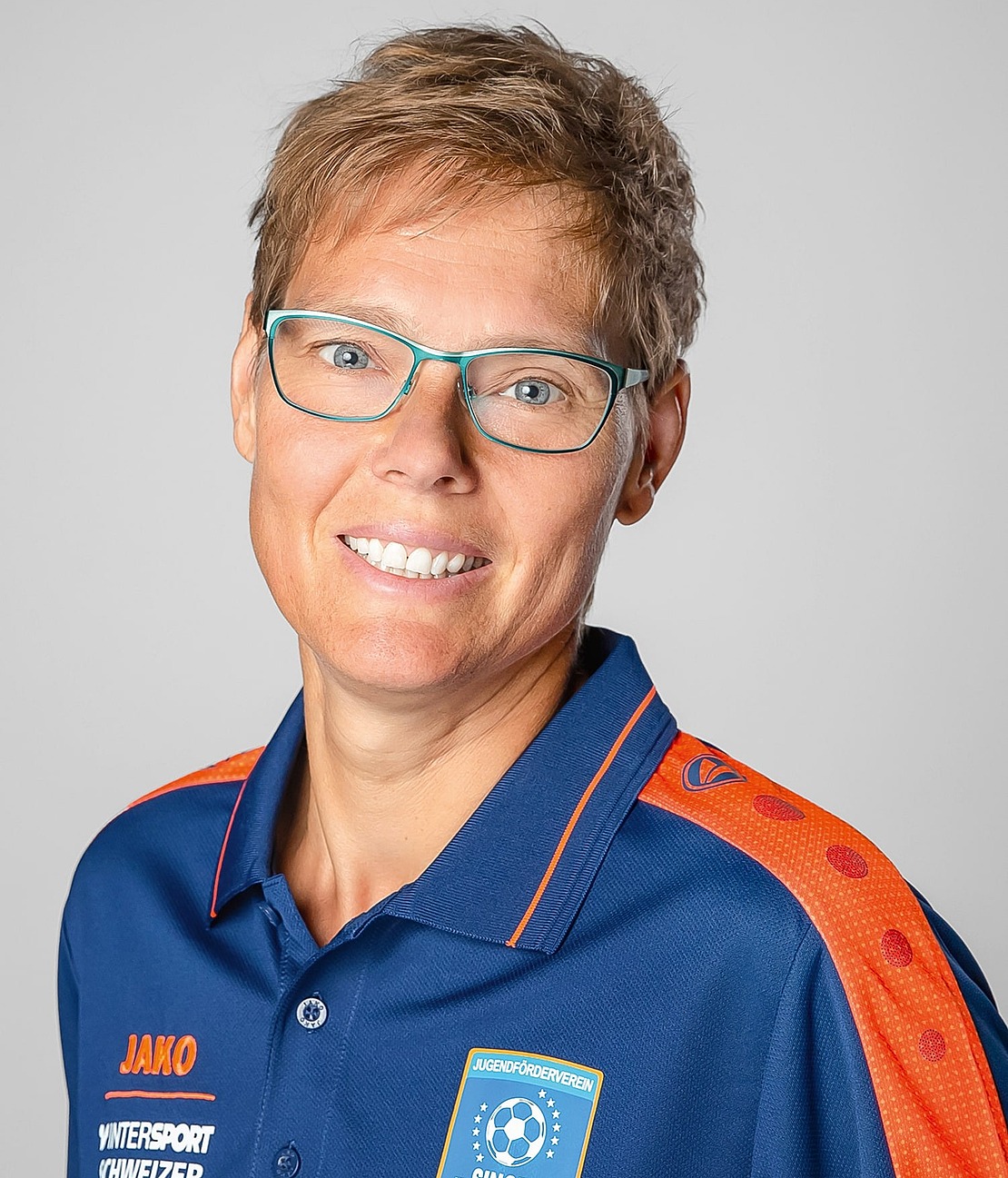 Eine Person mit kurzen hellbraunen Haaren und Brille, die ein blaues Sport-Poloshirt mit orangefarbenen Akzenten und Logos trägt, lächelt in die Kamera vor einem schlichten hellgrauen Hintergrund.