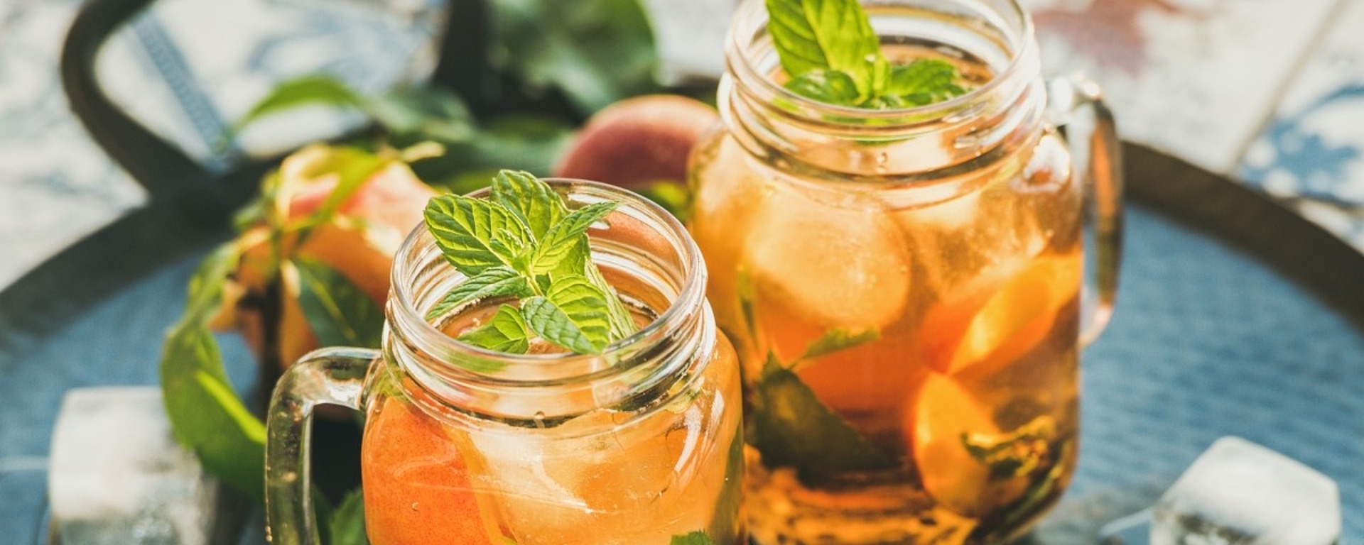 Zwei Einmachgläser, gefüllt mit Eistee, frischen Minzblättern und Orangenscheiben, stehen auf einem Tablett mit verstreuten Eiswürfeln und Minzezweigen auf einer gemusterten Oberfläche.