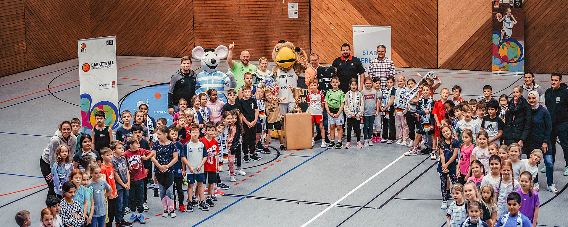 Eine große Gruppe von Kindern und Erwachsenen posiert gemeinsam in einer Turnhalle neben einem Basketballkorb. Im Hintergrund sind Transparente und zwei Personen in einem Maskottchenkostüm zu sehen. Viele der Kinder lächeln und blicken in die Kamera.