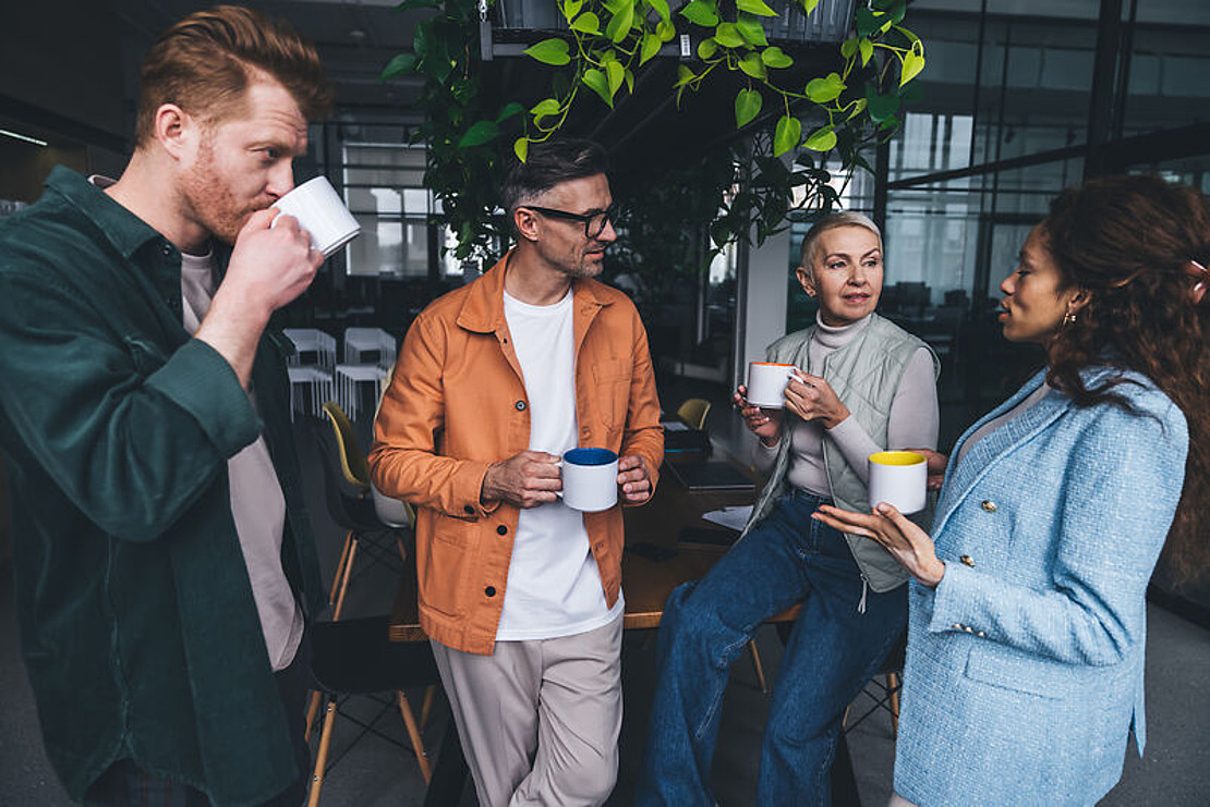 Vier Personen stehen und sitzen in einem Büro um einen Tisch herum, halten Tassen in der Hand und sind in ein Gespräch vertieft. Über ihnen stehen Pflanzen und im Hintergrund stehen Stühle. Die Atmosphäre wirkt entspannt und informell.