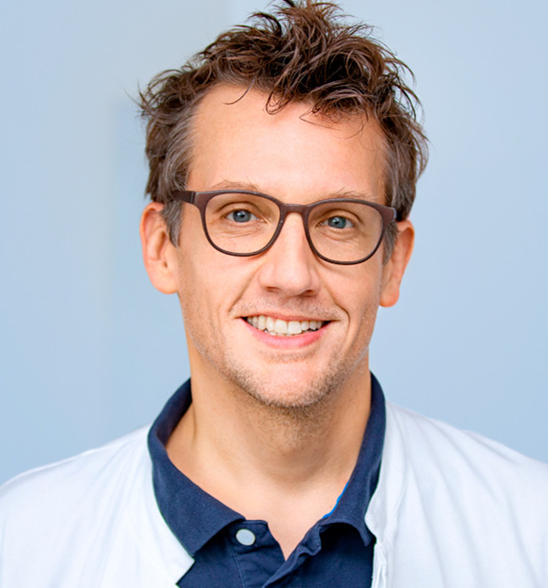Lächelnder Mann mit kurzen, zerzausten braunen Haaren und Brille, der ein marineblaues Poloshirt und einen weißen Mantel trägt, steht vor einem hellblauen Hintergrund.