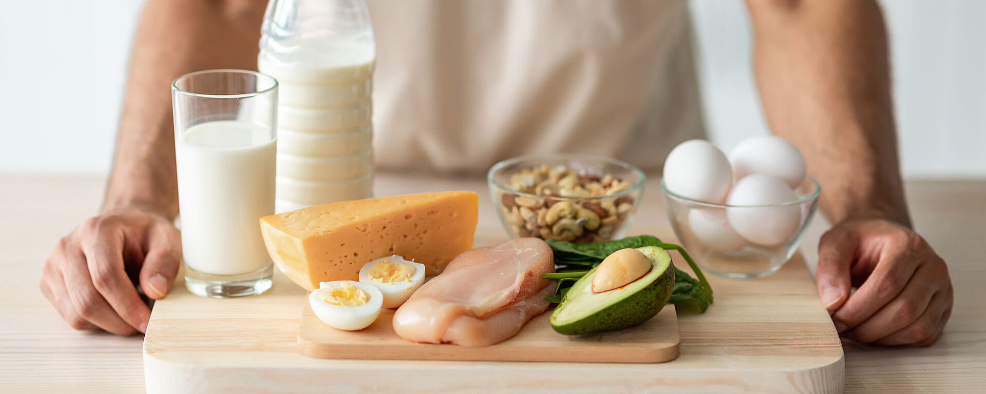 Eine Person steht hinter einem Schneidebrett, auf dem Eier, Avocado, Käse, Hähnchenbrust, Nüsse sowie Gläser und eine Flasche Milch ordentlich angeordnet sind.