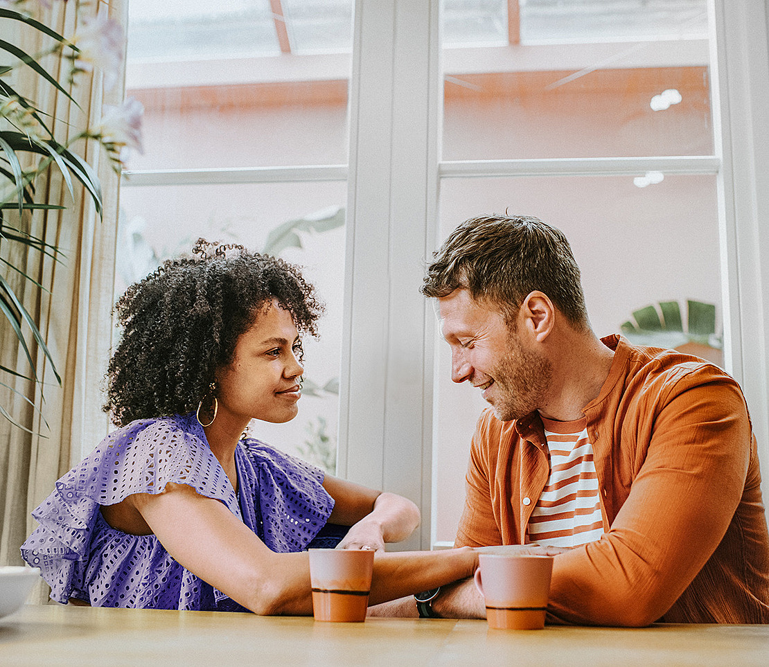 Eine Frau und ein Mann sitzen an einem Tisch in einem Haus, lächeln und schauen sich freundlich an. Jeder hat eine Tasse vor sich stehen, und die Umgebung ist hell und entspannt mit Pflanzen im Hintergrund.