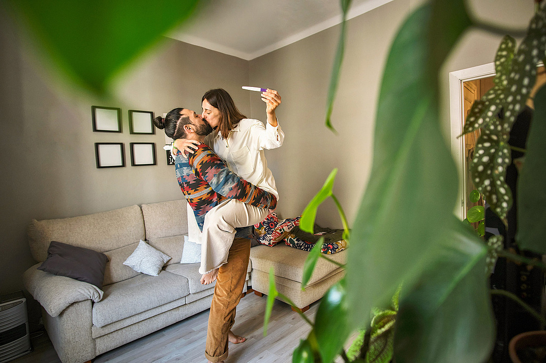 Ein glückliches Paar umarmt sich in einem gemütlichen Wohnzimmer. Die Frau mit einem positiven Schwangerschaftstest in der Hand wird von ihrem Partner hochgehoben, während sie feiern. Im Hintergrund stehen grüne Zimmerpflanzen und ein beiges Sofa.