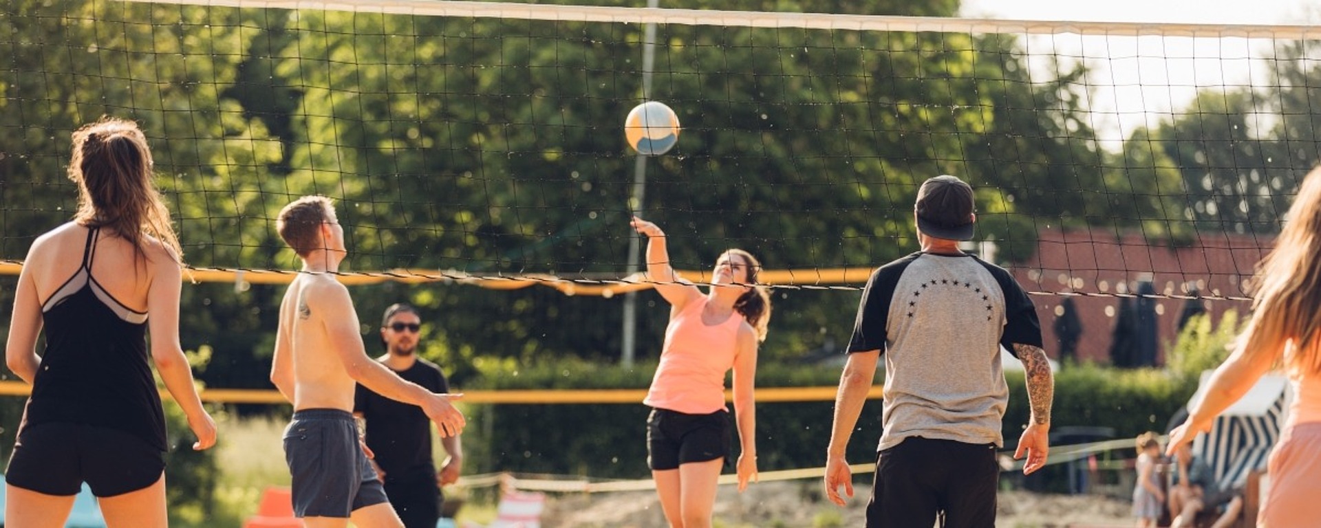 Sechs Personen spielen an einem sonnigen Tag Beachvolleyball. Eine Frau springt, um den Ball über das Netz zu schlagen, während die anderen zusehen oder sich darauf vorbereiten, zu reagieren. Im Hintergrund sind Bäume und Sitzgelegenheiten im Freien zu sehen.