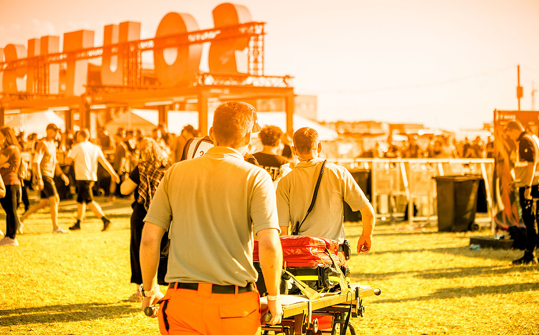 Zwei Rettungskräfte gehen mit einer leeren Trage durch ein überfülltes Open-Air-Festival. Die Szene ist hell mit einem warmen Orangefilter beleuchtet, im Hintergrund ist ein großes orangefarbenes Schild zu sehen.