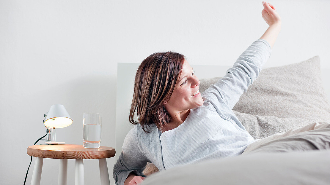 Eine Frau sitzt im Bett, lächelt und streckt ihren Arm aus. Neben ihr steht ein Nachttisch mit einem Glas Wasser und einer kleinen Lampe. Die Szene wirkt hell und gemütlich.