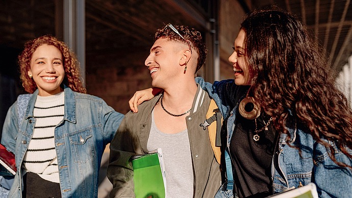 Drei junge Erwachsene, die lächelnd zusammen im Freien spazieren gehen und jeweils ein Notizbuch in der Hand halten. Zwei haben ihre Arme umeinander gelegt, und alle sind leger gekleidet, scheinen glücklich und in ein Gespräch vertieft zu sein.