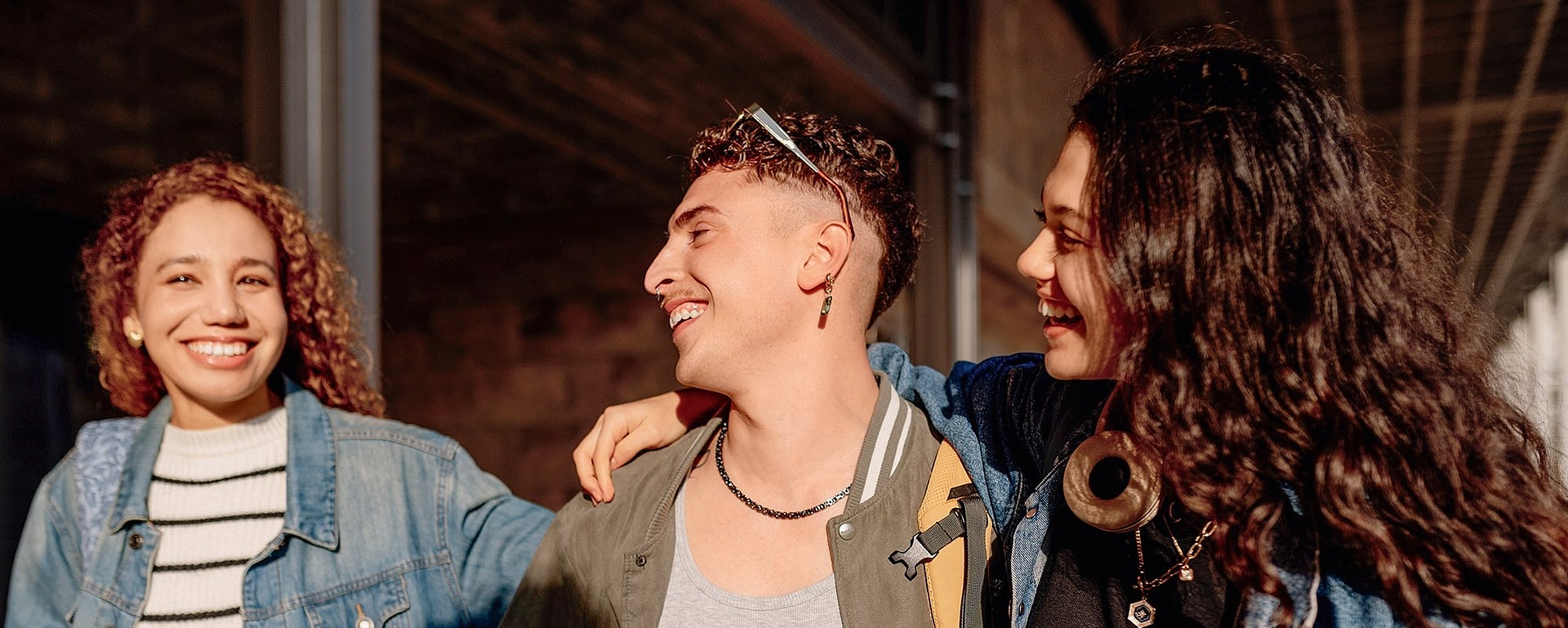 Drei junge Erwachsene, die lächelnd zusammen im Freien spazieren gehen und jeweils ein Notizbuch in der Hand halten. Zwei haben ihre Arme umeinander gelegt, und alle sind leger gekleidet, scheinen glücklich und in ein Gespräch vertieft zu sein.