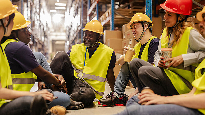 Eine Gruppe von Lagerarbeitern mit gelben Sicherheitswesten und Schutzhelmen sitzt im Kreis auf dem Boden, lächelt und plaudert, während sie in einem von Regalen und Kisten umgebenen Gang eine Kaffeepause einlegt.