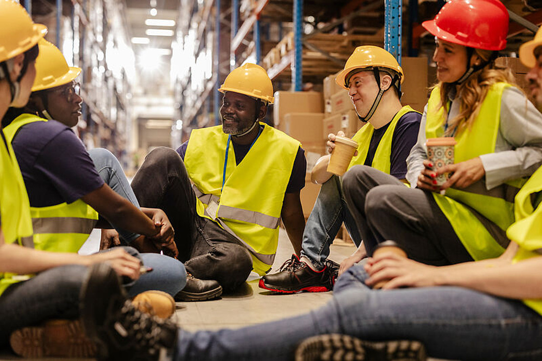 Eine Gruppe von Lagerarbeitern mit gelben Sicherheitswesten und Schutzhelmen sitzt im Kreis auf dem Boden, lächelt und plaudert, während sie in einem von Regalen und Kisten umgebenen Gang eine Kaffeepause einlegt.