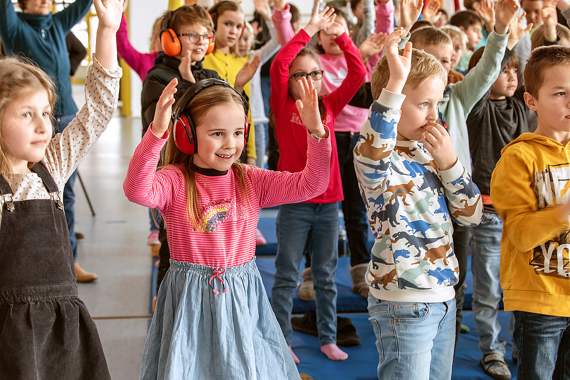 Eine Gruppe kleiner Kinder steht in einem Haus dicht beieinander, hebt die Arme und lächelt. Ein Mädchen im Vordergrund trägt pinke Kopfhörer, und andere tragen bunte Kleidung, während sie an einer Aktivität teilnehmen.