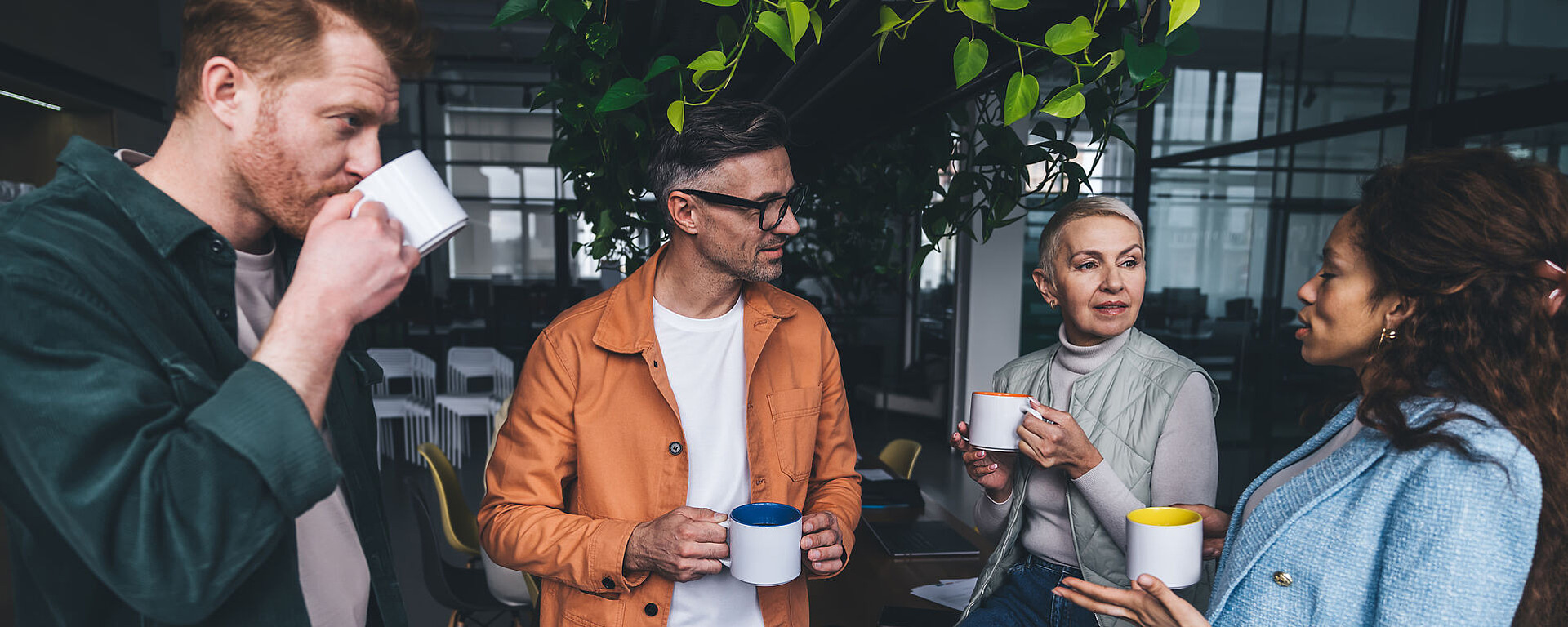 Vier Personen stehen in einem Innenraum zusammen, halten Tassen in der Hand und führen ein Gespräch. Zwei Männer und zwei Frauen, alle in Freizeit- oder Geschäftskleidung gekleidet, mit Pflanzen und Büromöbeln im Hintergrund.