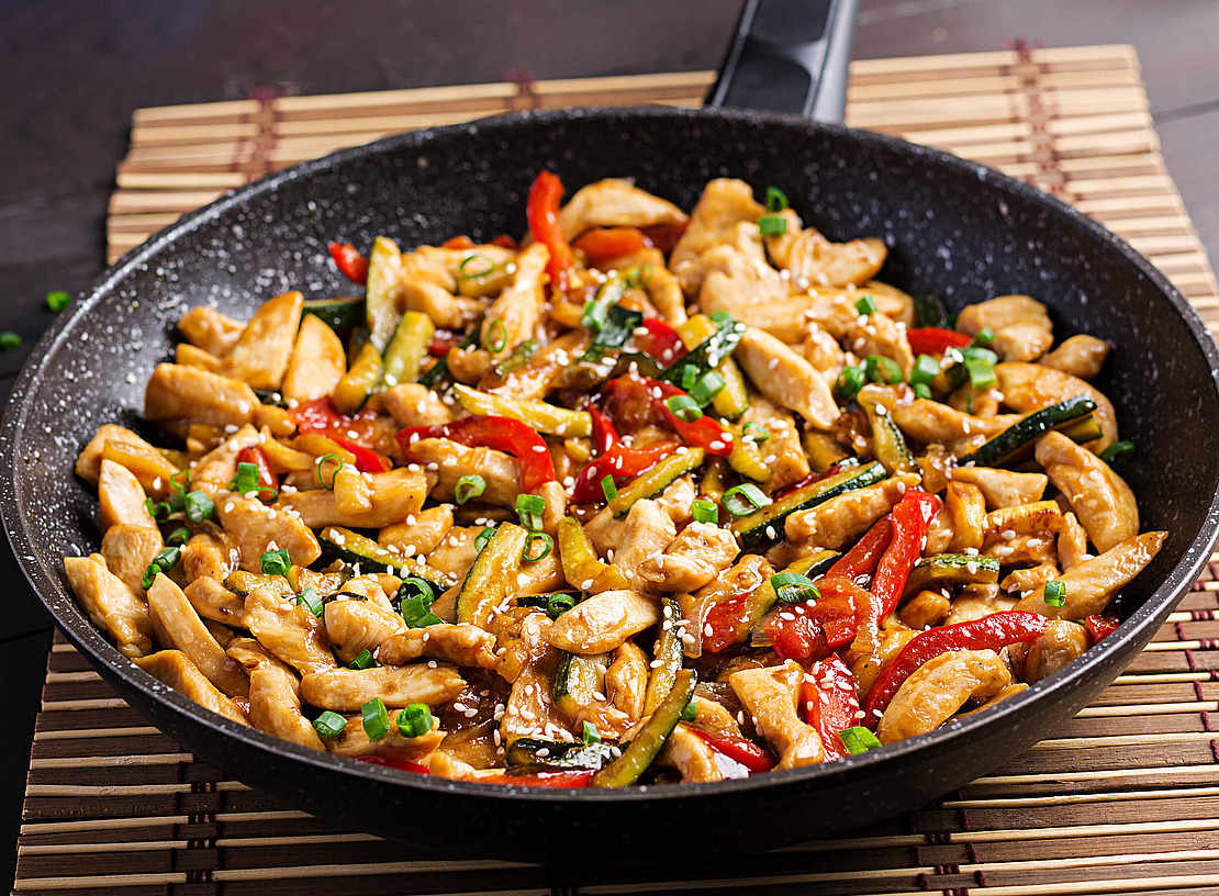 Eine schwarze Bratpfanne gefüllt mit gebratenen Hähnchenstreifen, roten Paprikaschoten, Zucchini und Frühlingszwiebeln, garniert mit Sesamkörnern, auf einem Tischset aus Bambus.