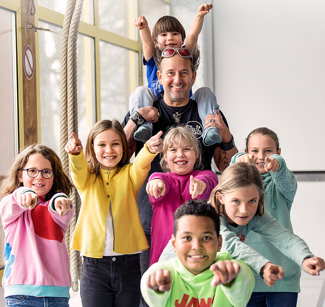Ein lächelnder Erwachsener mit einem Kind auf den Schultern und fünf weitere lächelnde Kinder stehen in einem Haus und zeigen aufgeregt in Richtung der Kamera. Die Gruppe ist fröhlich und energiegeladen, mit hellem Tageslicht im Hintergrund.