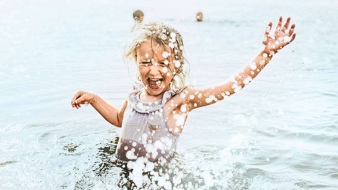 Ein kleines Kind lächelt freudig mit einem erhobenen Arm und spritzt Wasser, während es in einem flachen, sonnenbeschienenen Gewässer steht. Im Hintergrund sind Wassertropfen und zwei verschwommene Figuren zu sehen.