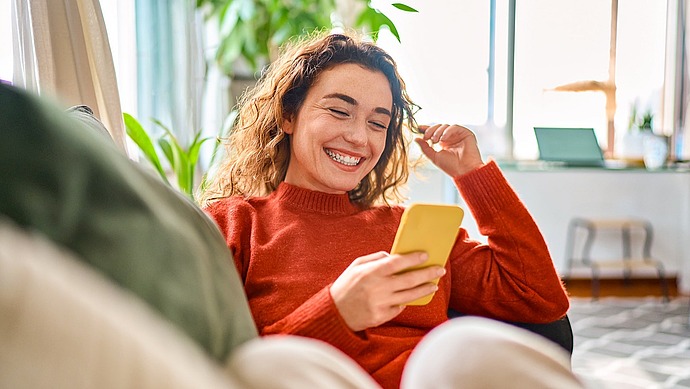 Eine junge Frau mit lockigem Haar und Zahnspange, die einen roten Pullover trägt, lächelt, während sie auf ihr gelbes Smartphone schaut. Sie sitzt gemütlich in einem Raum mit Pflanzen und Sonnenlicht im Hintergrund.