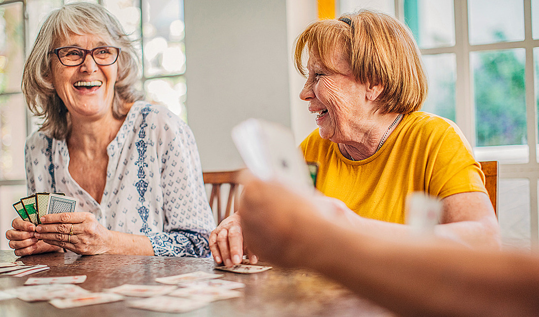 Zwei ältere Frauen sitzen an einem Tisch, lächeln und lachen, während sie mit anderen Karten spielen. Das Sonnenlicht fällt durch die großen Fenster hinter ihnen ein und schafft eine warme und fröhliche Atmosphäre.