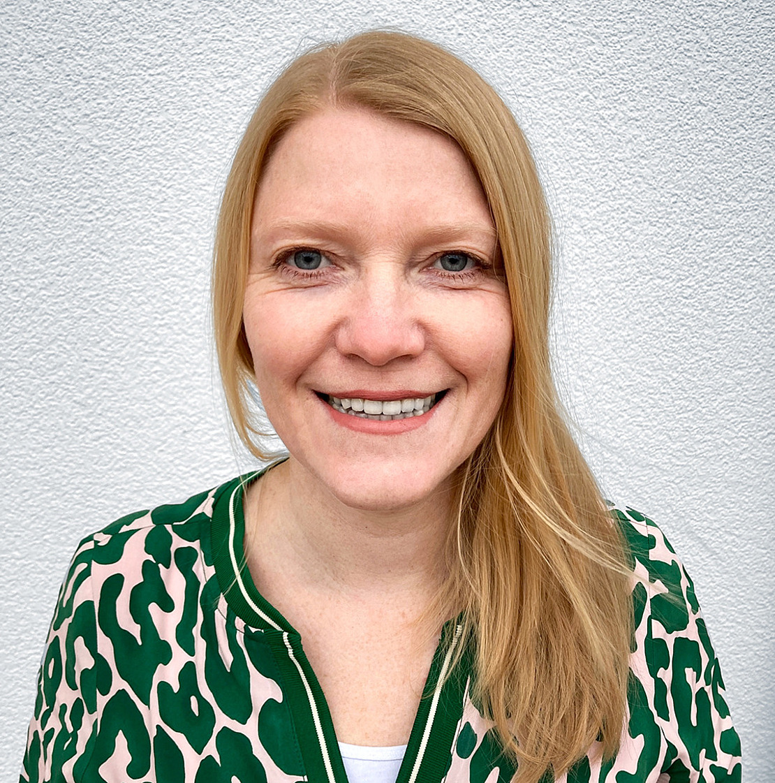 Eine Frau mit glattem blondem Haar und blauen Augen lächelt in die Kamera. Sie trägt eine grüne und beigefarbene Bluse mit Tiermuster und steht vor einer weißen, strukturierten Wand.