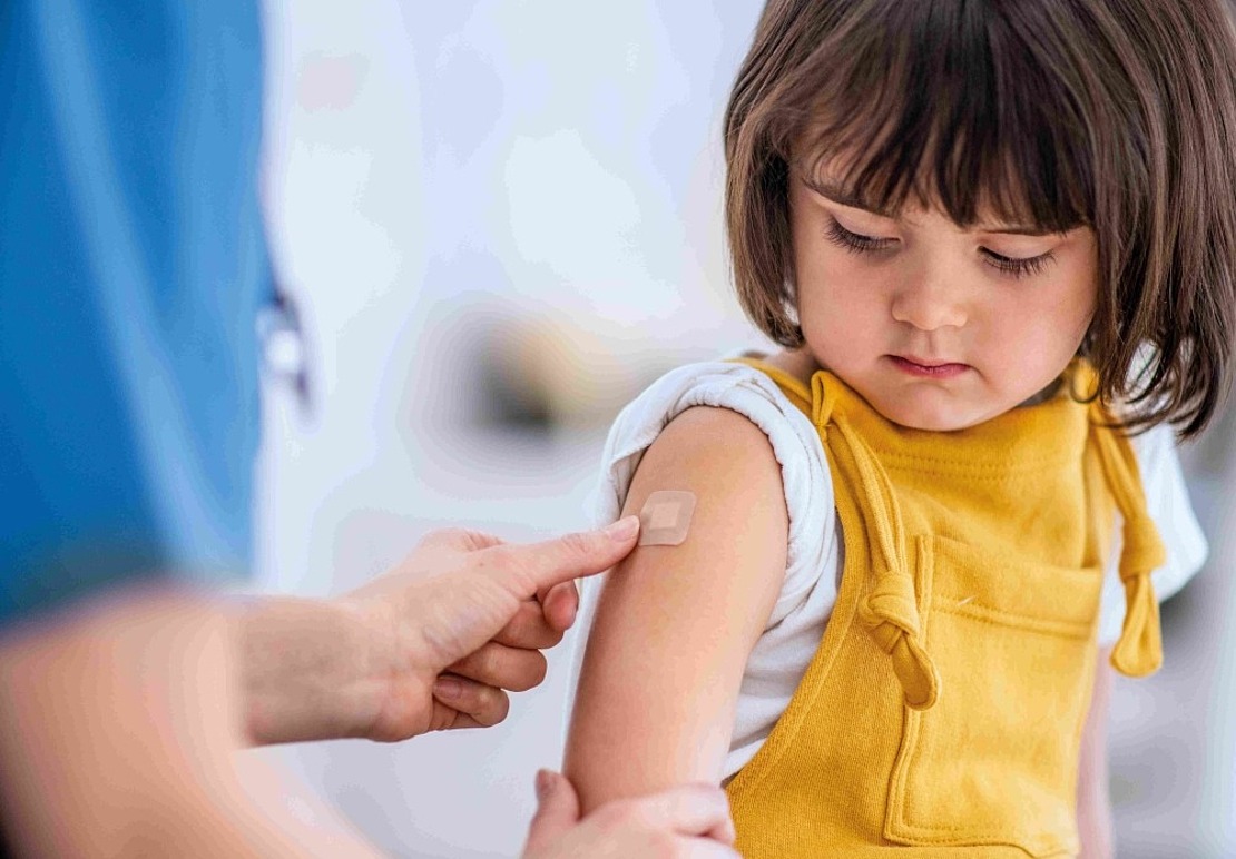 Ein kleines Kind in gelber Kleidung betrachtet einen kleinen Pflasterverband, der ihm von einem Erwachsenen auf den Oberarm geklebt wird, wahrscheinlich nach einer Impfung oder Injektion.