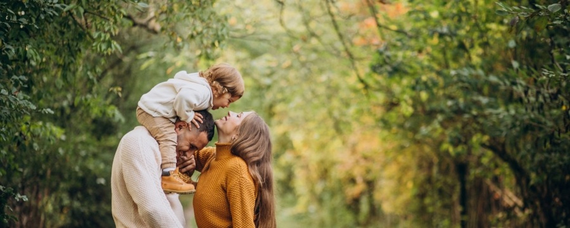 Eine vierköpfige Familie steht auf einem belaubten Waldweg. Ein Mann hält ein kleines Kind auf seinen Schultern, das sich herunterbeugt, um eine Frau zu küssen. Ein anderes Kind steht davor, lächelt und hält die Hand der Frau. Sie sind von Bäumen umgeben.