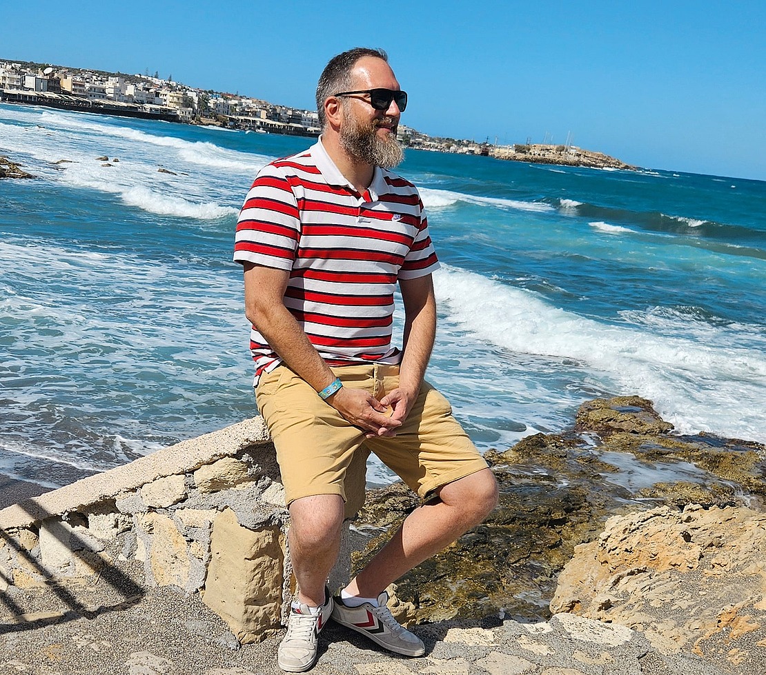 Ein Mann mit Bart, der eine Sonnenbrille, ein rot-weiß gestreiftes Hemd und hellbraune Shorts trägt, sitzt auf einer Steinmauer am Meer, während die Wellen an die Felsen schlagen und eine Küstenstadt im Hintergrund unter einem klaren blauen Himmel zu sehen ist.