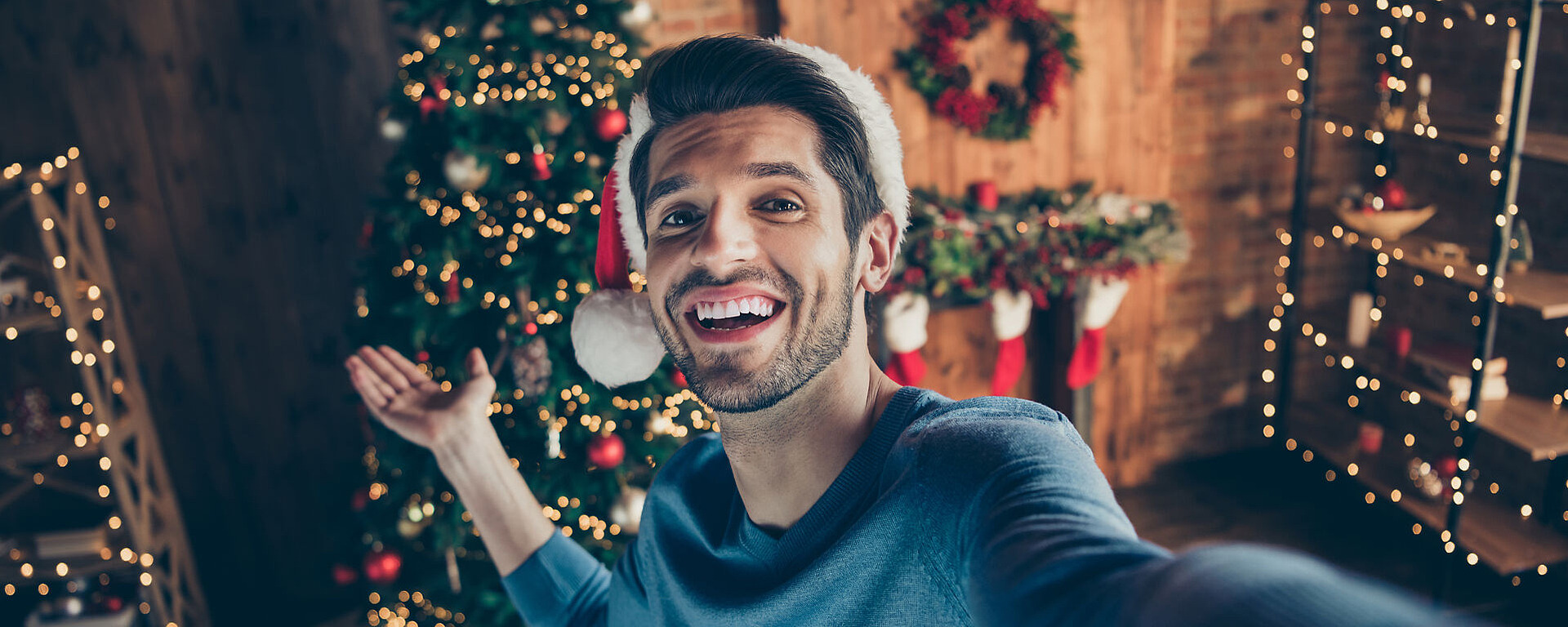 Eine lächelnde Person mit einer Weihnachtsmannmütze macht ein Selfie vor einem geschmückten Weihnachtsbaum mit eingepackten Geschenken, Strümpfen und festlichen Lichtern in einem gemütlichen Raum.