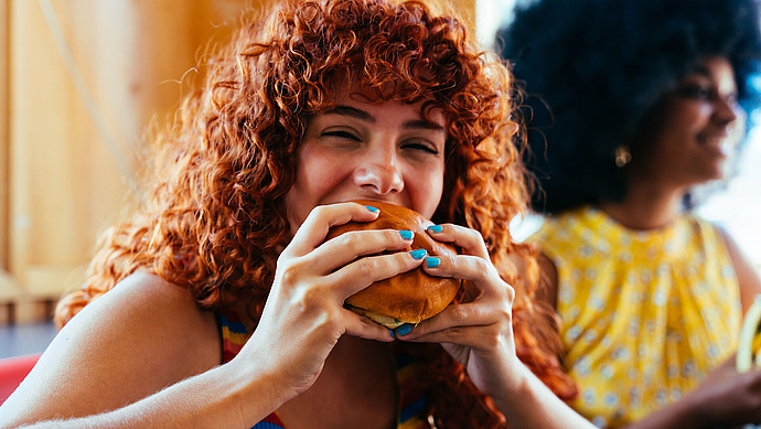 Eine Person mit lagen lockigem rotem Haar und leuchtend blauem Nagellack isst vergnügt einen Burger. Eine weitere Person mit lockigem schwarzem Haar ist im Hintergrund teilweise sichtbar und trägt ein gelb gemustertes Oberteil. Die Umgebung wirkt hell und leger.