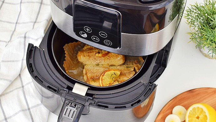 Ein Frittierkorb mit gegarten Fischfilets, garniert mit Zitronenscheiben, neben einem Teller mit weiteren Zitronenscheiben und Kräutern auf einer weißen Arbeitsplatte.