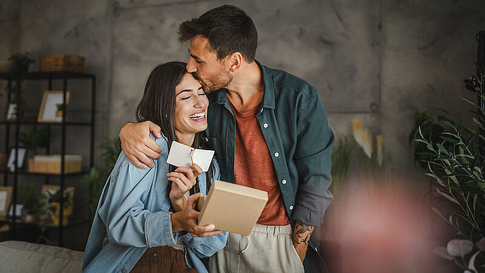 Ein Mann umarmt und küsst eine lächelnde Frau auf den Kopf, während sie ein Geschenkpaket und eine Karte hält. Sie wirken glücklich und stehen in einem gemütlichen, modernen Wohnzimmer mit Regalen und Pflanzen im Hintergrund.