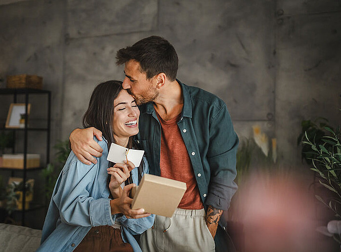Ein Mann umarmt und küsst eine lächelnde Frau auf den Kopf, während sie ein Geschenkpaket und eine Karte hält. Sie wirken glücklich und stehen in einem gemütlichen, modernen Wohnzimmer mit Regalen und Pflanzen im Hintergrund.