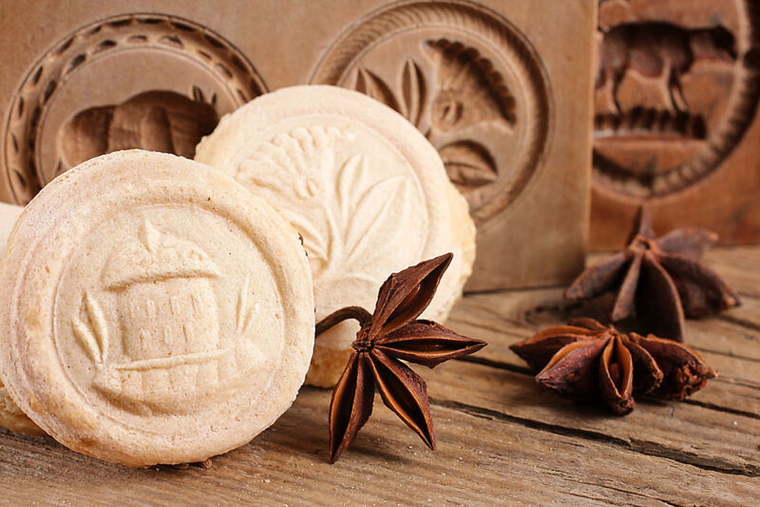 Auf einem Holztisch liegen runde Kekse mit aufwändig gestanzten Mustern, begleitet von Sternanis. Im Hintergrund sind Holzformen mit ähnlichen Mustern zu sehen. Die Szene erinnert an eine rustikale, traditionelle Backatmosphäre.