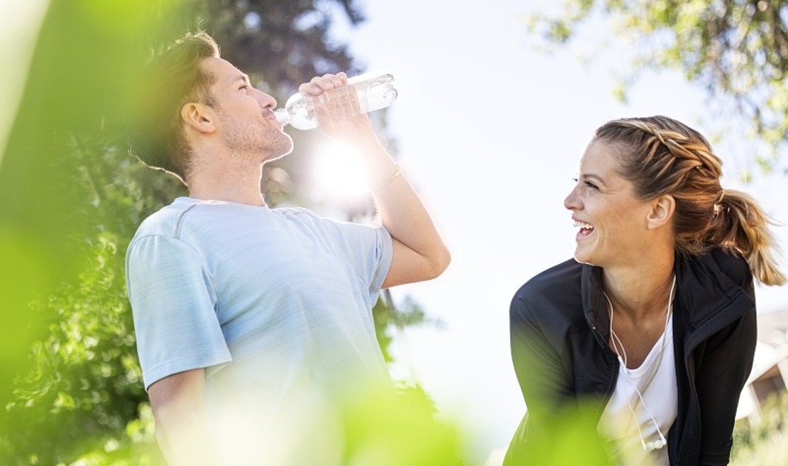 Ein Mann trinkt Wasser aus einer Flasche, während eine Frau neben ihm lächelt und lacht. Beide sind sportlich gekleidet und genießen eine sonnige Umgebung im Freien mit grünem Laub im Vordergrund.
