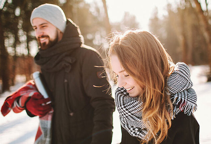 Eine junge Frau und ein Mann in Winterkleidung gehen an einem verschneiten Tag gemeinsam im Freien spazieren. Die Frau lächelt, und das Sonnenlicht fällt durch die Bäume im Hintergrund.