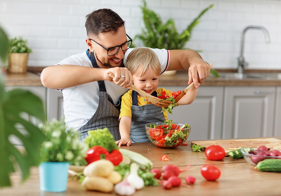 Ein Mann und ein kleines Kind, die beide Schürzen tragen, bereiten gemeinsam einen frischen Salat in einer Küche zu. Sie lächeln und benutzen hölzerne Utensilien, um Gemüse in einer Glasschüssel auf einer Holztheke zu mischen.