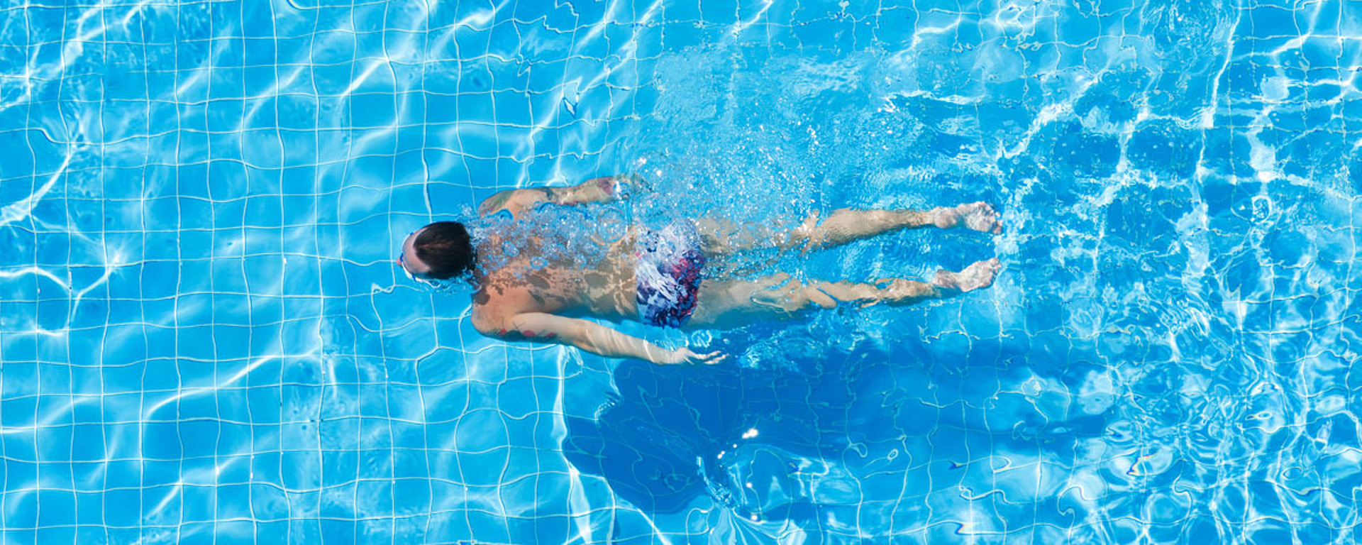 Eine Person schwimmt allein im Freistil in einem hellblauen Schwimmbecken. Das Sonnenlicht wird vom Wasser reflektiert, wodurch schimmernde Muster entstehen. In der oberen rechten Ecke steht in einer Sprechblase "MACH ES!" mit einem blauen Herz.
