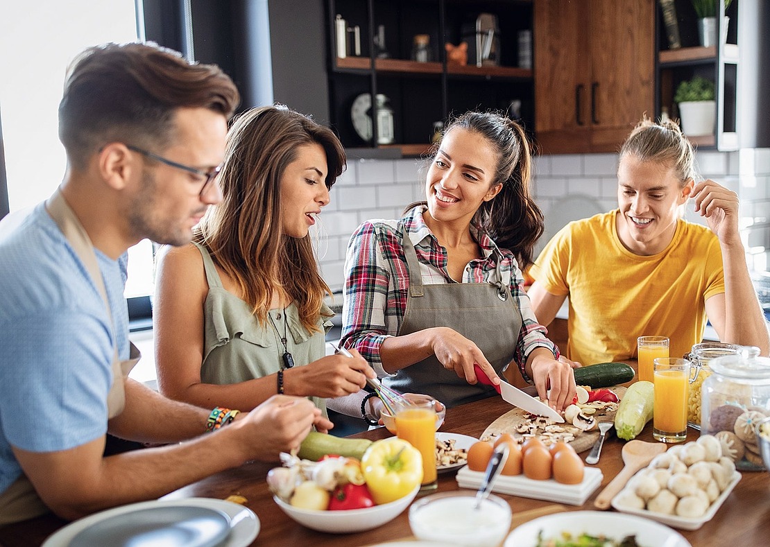 Vier junge Erwachsene sitzen zusammen in einer Küche, bereiten Gemüse und andere Zutaten auf einem Holztisch zu, lächeln und genießen die Gesellschaft des anderen.