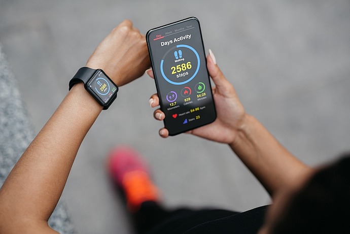 Eine Person schaut auf ein Smartphone, auf dem eine Fitness-App mit Aktivitätsstatistiken angezeigt wird, die 2.586 Schritte anzeigt. Sie trägt auch eine Smartwatch, und im Hintergrund ist ihr hellrosa Schuh zu sehen.