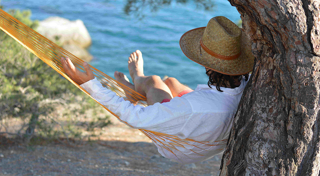 Eine Person, die einen Strohhut und ein weißes Hemd trägt, entspannt sich an einem sonnigen Tag in einer gelben Hängematte im Schatten, die an einem Baum festgebunden ist, mit hochgelegten Beinen und Blick auf ein blaues Gewässer.