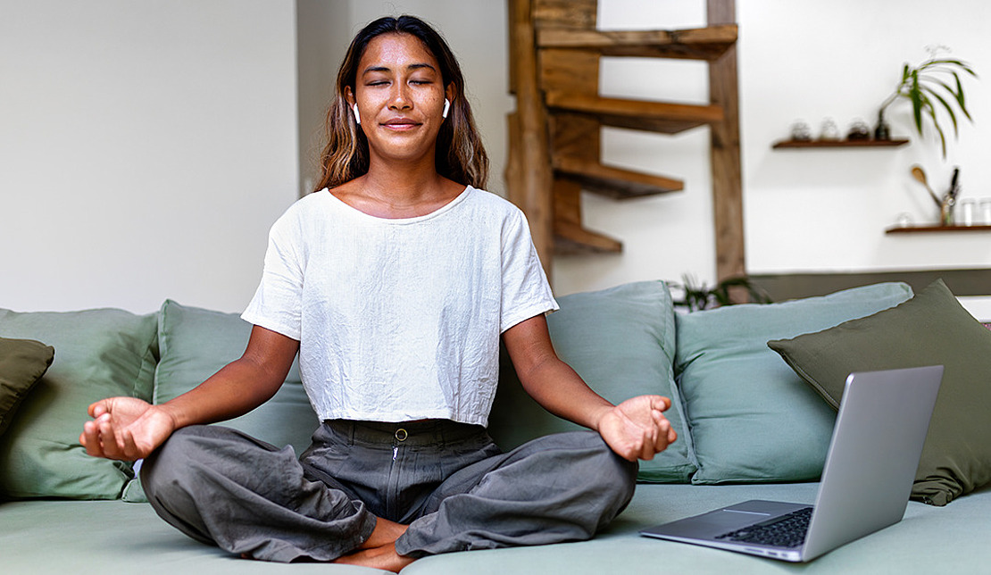 Eine Person sitzt im Schneidersitz auf einem Sofa und meditiert mit geschlossenen Augen und Ohrstöpseln. In der Nähe steht ein geöffneter Laptop. Die Szenerie ist ein gemütlicher Raum mit einer hölzernen Wendeltreppe und minimalistischem Dekor.