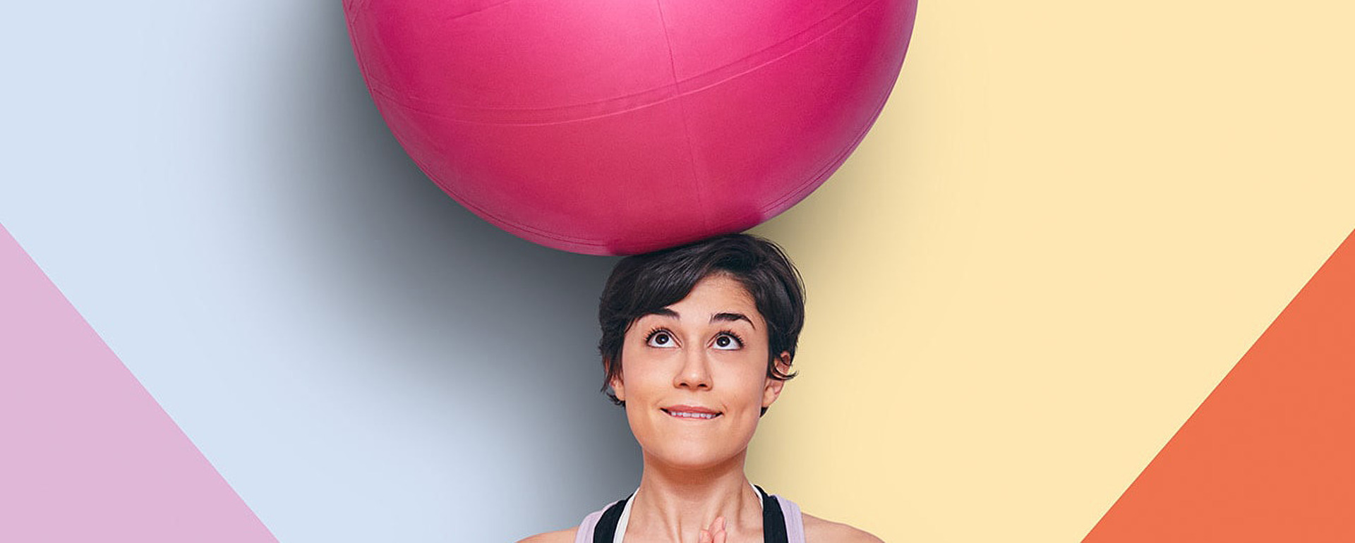 Eine Frau sitzt im Schneidersitz auf einer Yogamatte, hält die Hände in Gebetshaltung und balanciert einen großen rosafarbenen Gymnastikball auf ihrem Kopf. Im Hintergrund sind bunte geometrische Formen zu sehen.