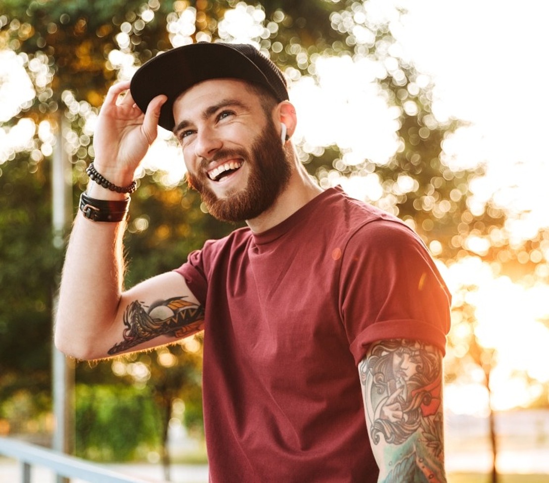 Ein tätowierter Mann, der ein rotes T-Shirt und eine rote Mütze trägt, lächelt in einer sonnigen Umgebung im Freien.