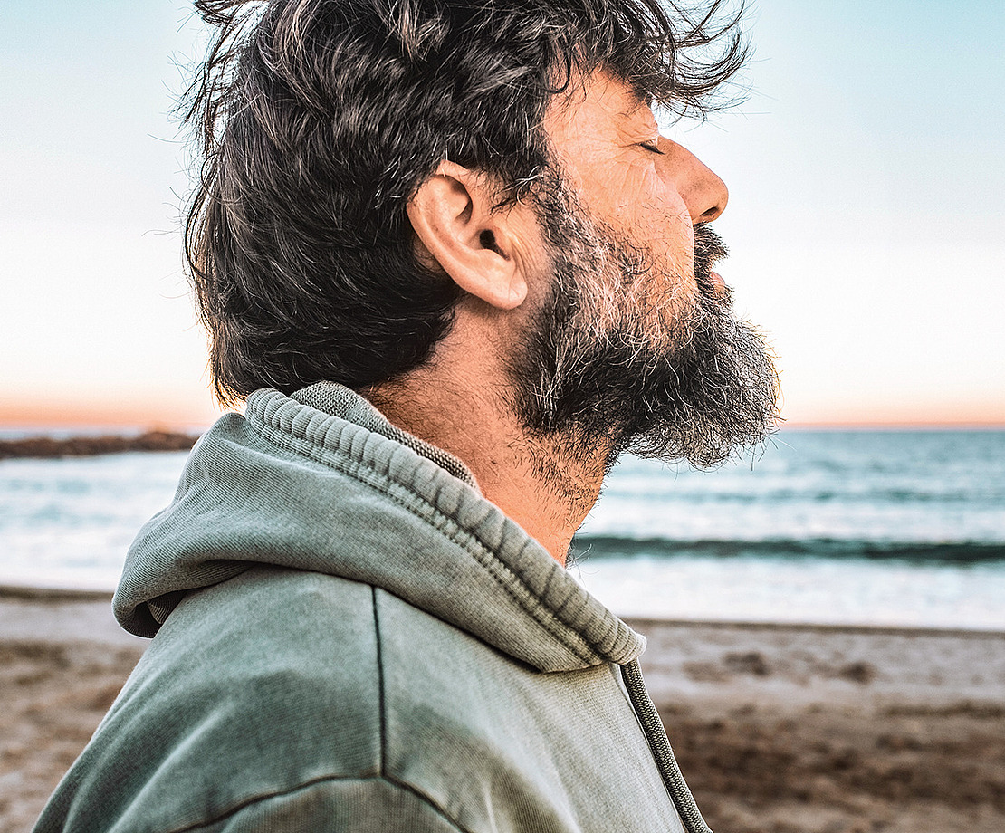 Ein bärtiger Mann in einem grünen Kapuzenpulli steht mit geschlossenen Augen an einem Strand und blickt aufs Meer. Der Himmel ist klar, und im Hintergrund sind das Meer und der Sandstrand zu sehen.