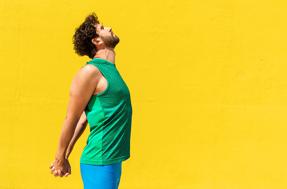 Ein Mann in einem grünen ärmellosen Hemd und einer blauen Hose macht eine Brustmuskel-Dehnübung.