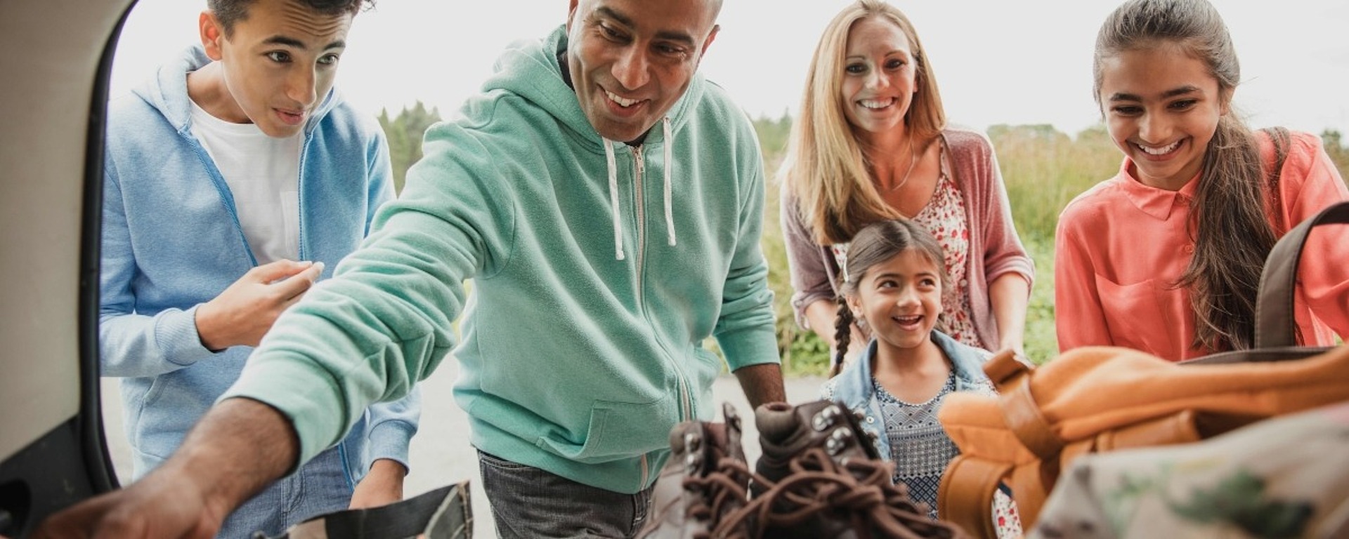 Eine fünfköpfige Familie, darunter zwei Erwachsene und drei Kinder, die lächelnd Gegenstände wie Stiefel und Taschen aus dem Kofferraum eines Autos auslädt, um sich auf eine Aktivität oder ein Abenteuer im Freien vorzubereiten.