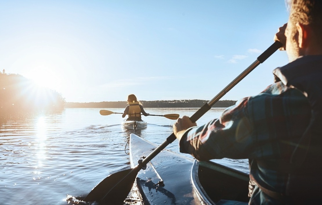 Zwei Kajakfahrer auf einem ruhigen See bei Sonnenaufgang oder Sonnenuntergang. Die Person im Vordergrund paddelt und folgt dem zweiten Kajakfahrer, wobei sich das Sonnenlicht auf dem Wasser spiegelt und die Bäume in der Ferne zu sehen sind.