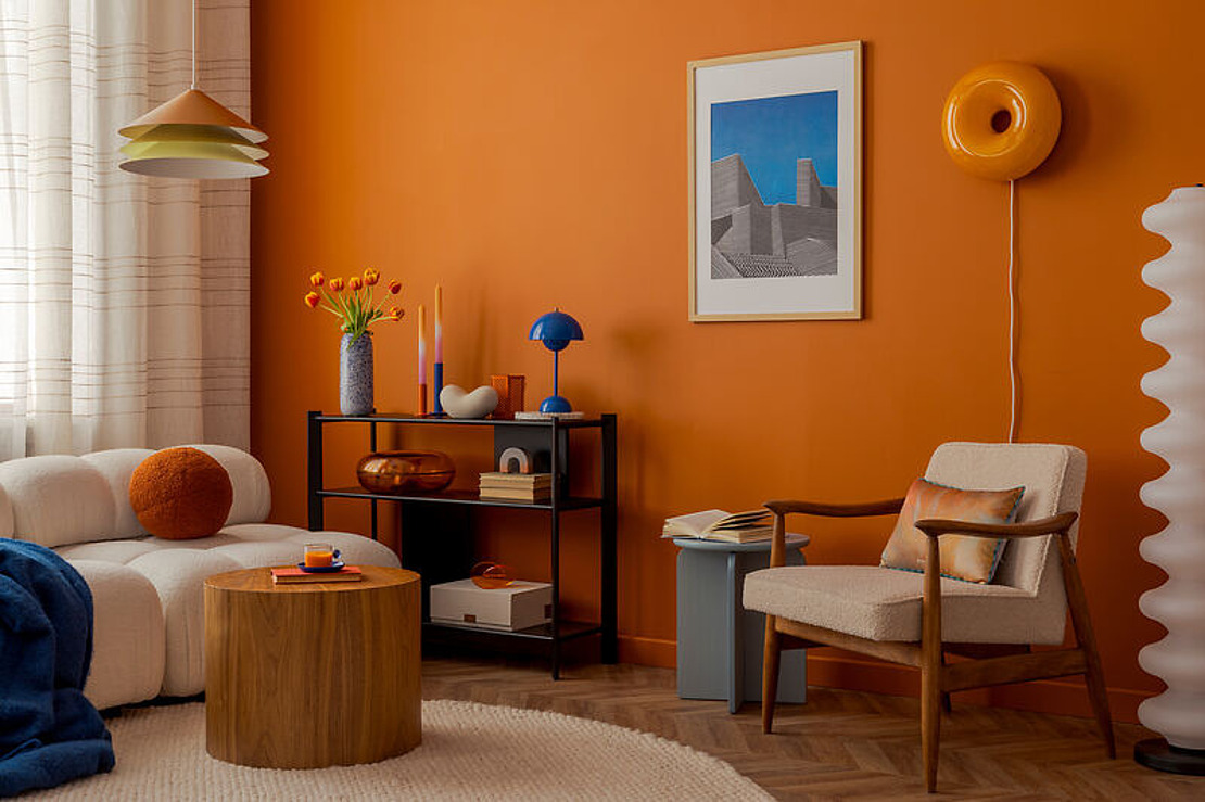 Ein modernes Wohnzimmer mit orangefarbenen Wänden, Mid-Century-Möbeln, einem weißen Sofa mit orangefarbenem Kissen, einem runden Couchtisch aus Holz, Kunstwerken und Dekorationsartikeln in Blau- und Orangetönen. Eine Stehlampe und Regale vervollständigen den gemütlichen Raum.