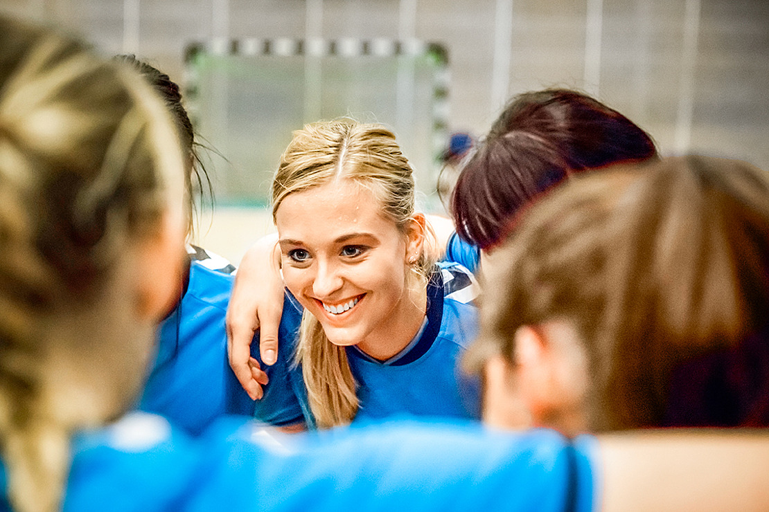 Eine junge Frau in einem blauen Sportdress lächelt, während sie sich mit ihren Mannschaftskameraden in einer Turnhalle zusammensetzt, was auf Teamgeist und Kameradschaft vor einem Spiel schließen lässt.