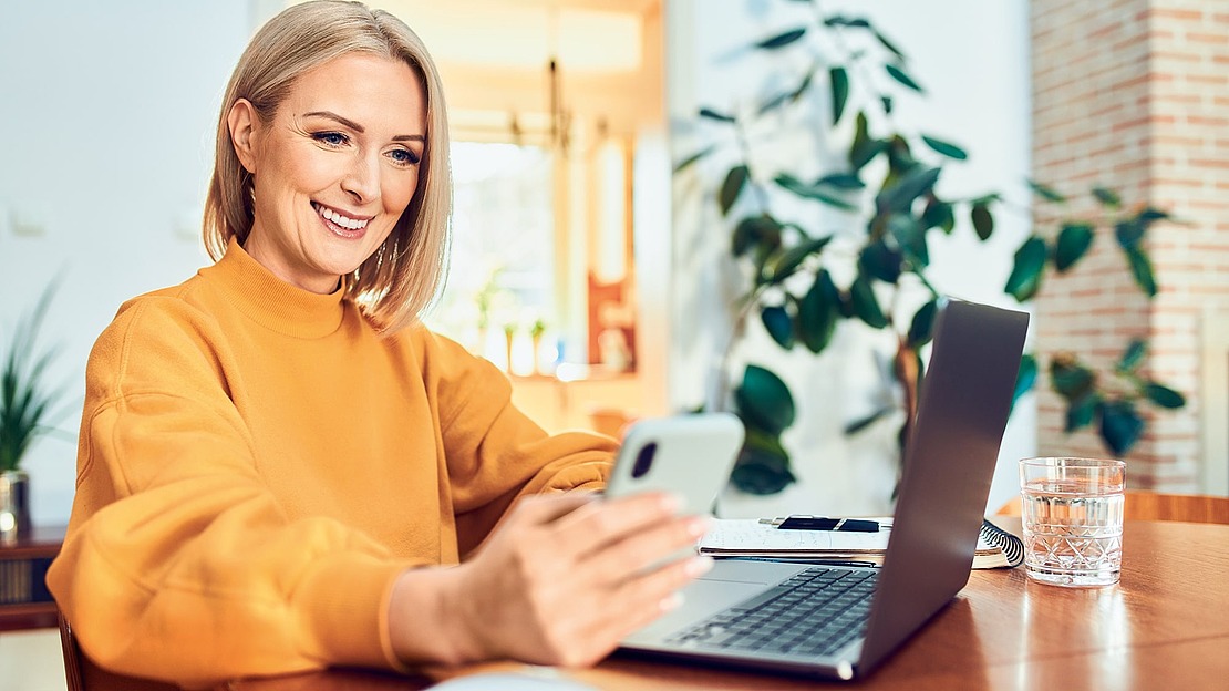 Eine lächelnde Frau in einem orangefarbenen Pullover sitzt an einem Schreibtisch mit einem Laptop und hält ein Smartphone in der Hand. Ein Glas Wasser, eine Maus und eine Topfpflanze stehen in der Nähe in einem hellen, gemütlichen Raum.